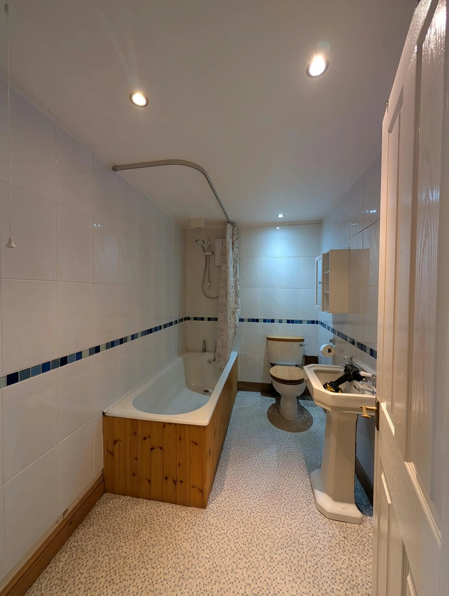 Existing narrow bathroom with white tiles, a blue mosaic border and a timber bath panel.