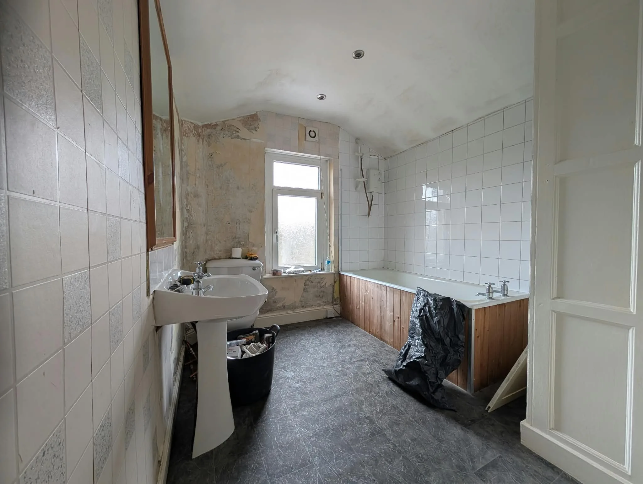 Original bathroom viewed from the doorway, with a bath, basin and worn wall finishes.